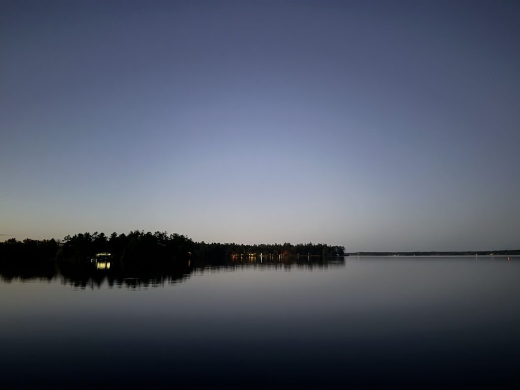 Still lake under a night sky with faint stars and distant shoreline lights reflecting on the water