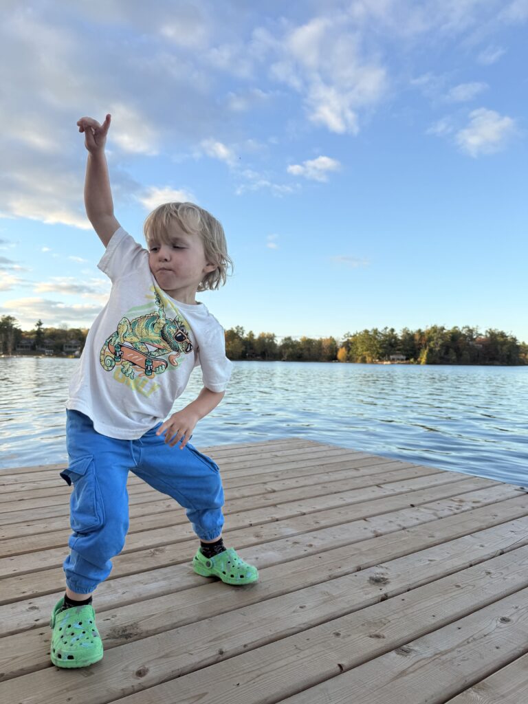Young child posing playfully on a wooden dock by a calm lake under a blue sky with scattered clouds