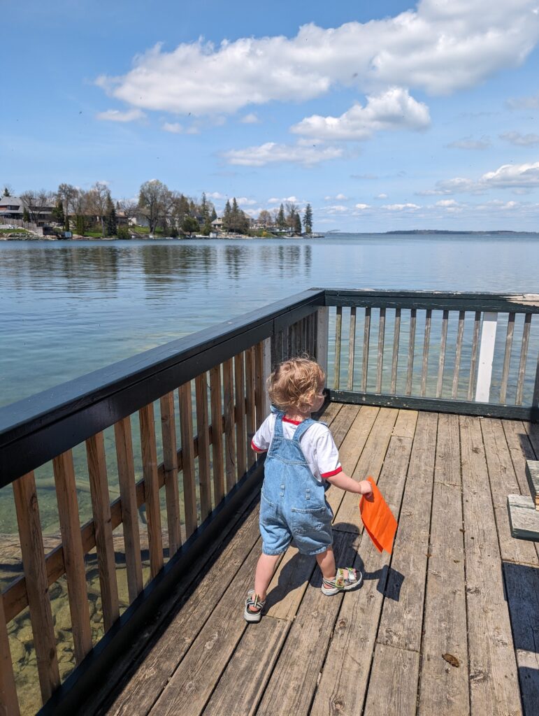 Young child walking along a wooden lakeside deck holding an orange toy with clear water and shoreline in the background