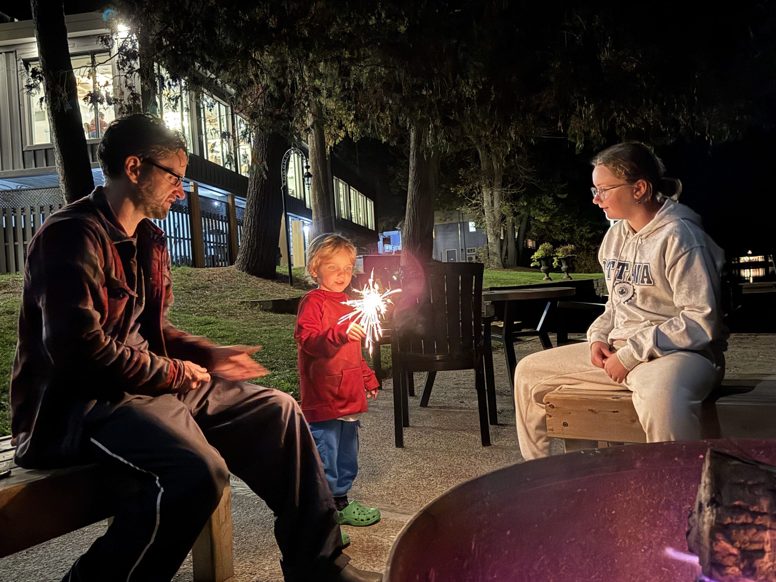Child holding a lit sparkler while sitting with a teenager and adult around a campfire at night beside a lakeside patio