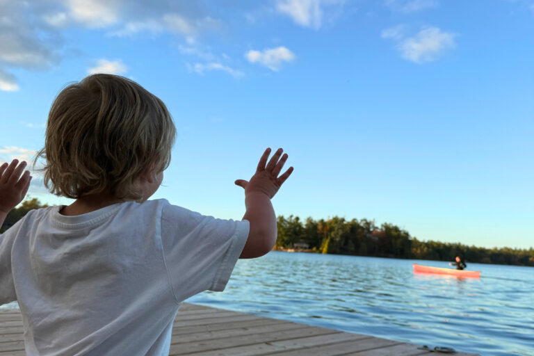 A young child standing on a dock, waving toward a red canoe on a calm lake under a bright blue sky with trees along the shoreline.