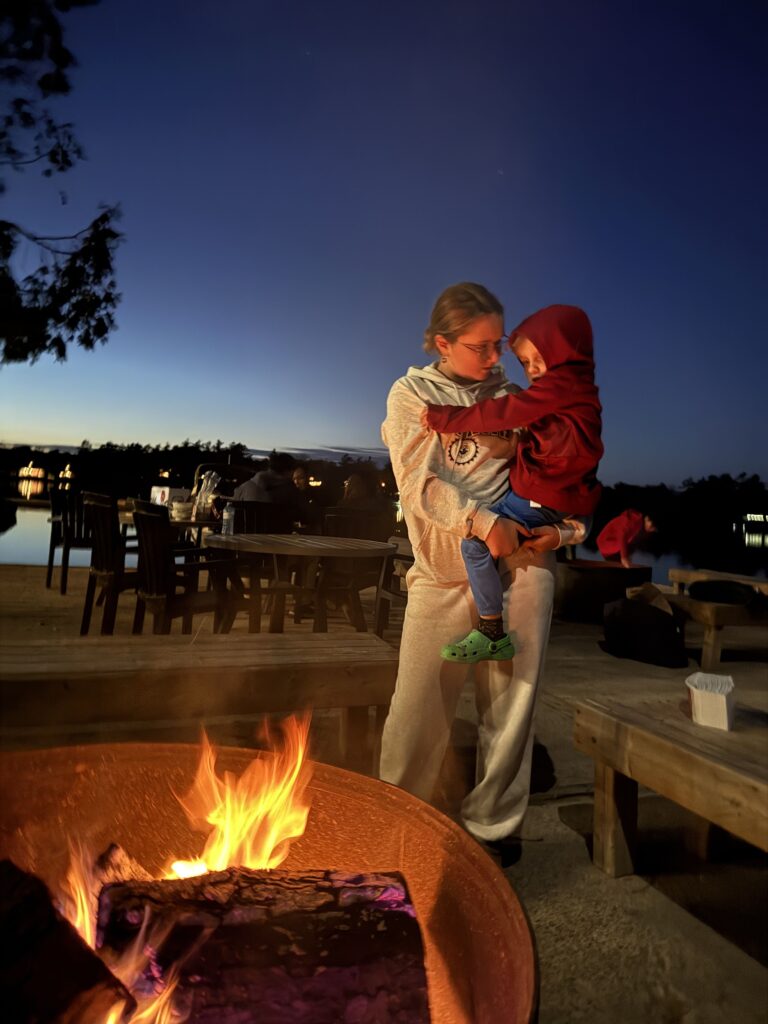 Teenager sibling holding younger child near a glowing campfire at dusk with outdoor seating and lake visible in the background