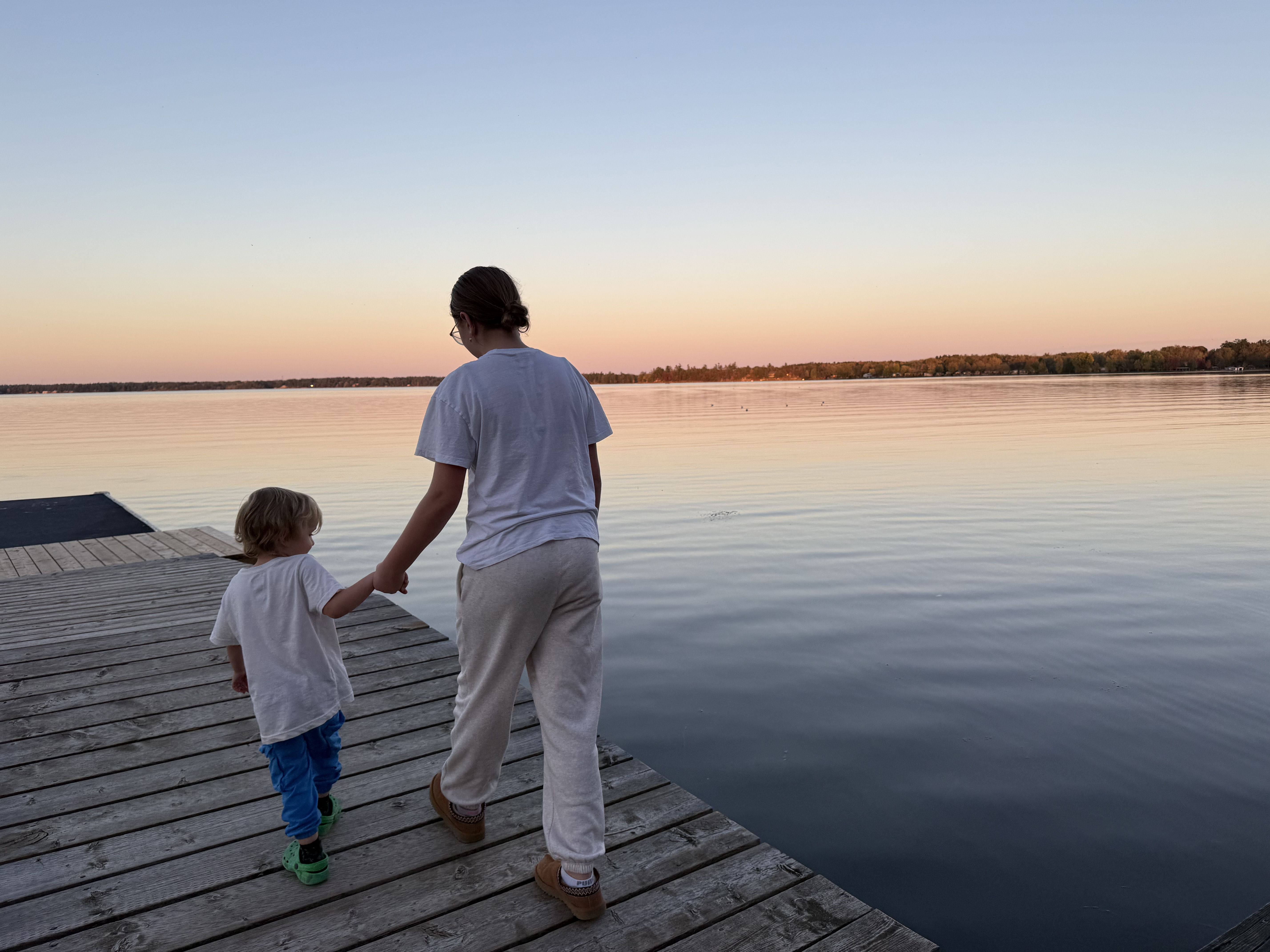 Teenager and child holding hands while walking along a wooden dock beside a calm lake during sunset