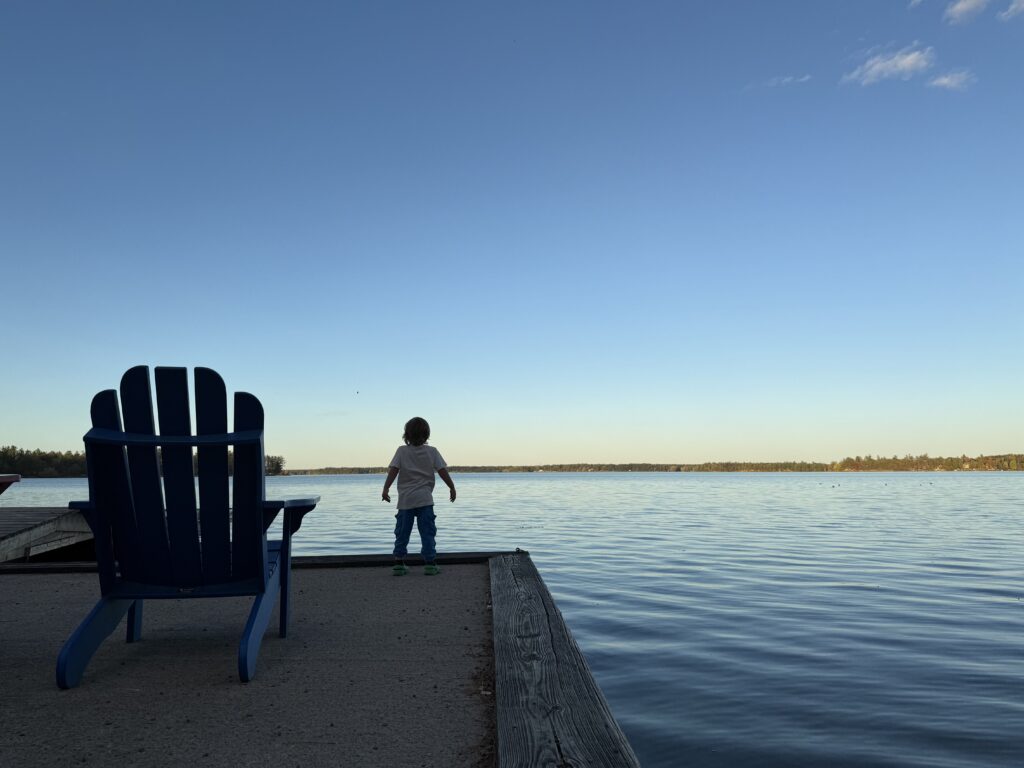 Child standing at the edge of a lakeside dock beside a blue chair, looking out over calm water with a distant tree-lined shoreline and open sky