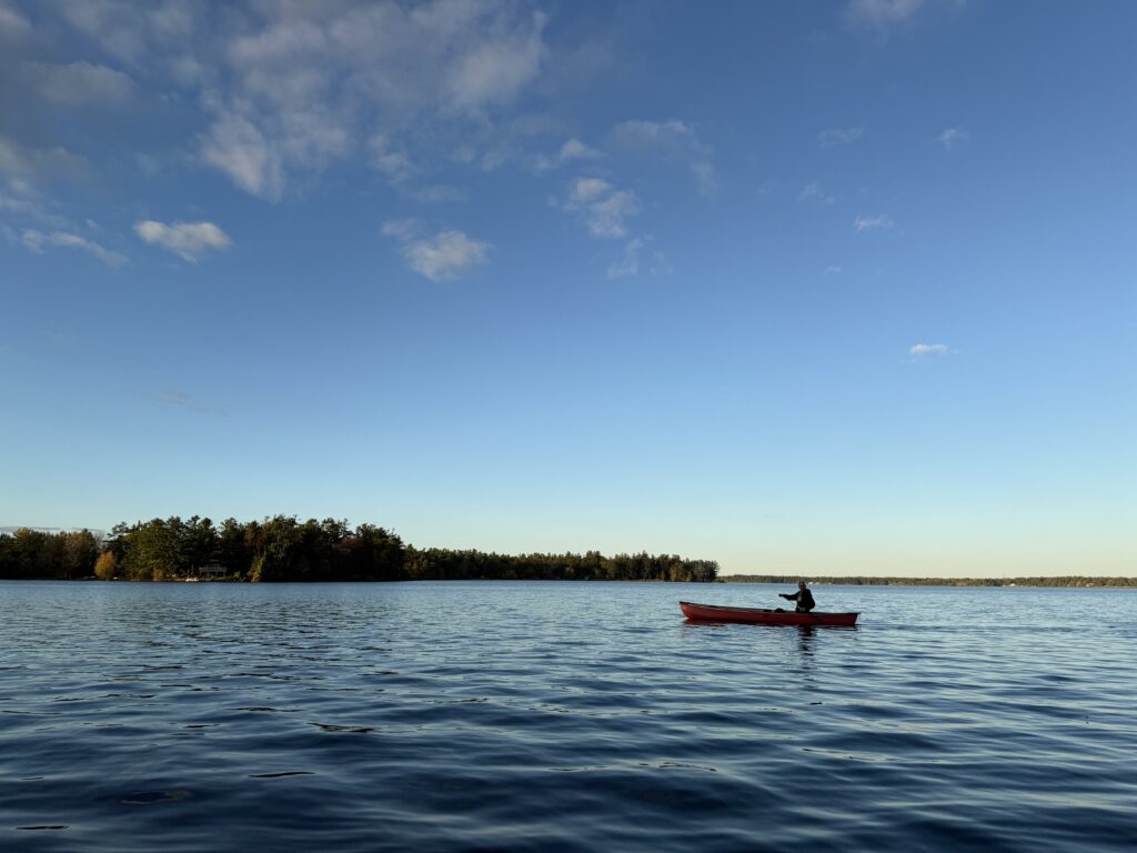 A red canoe moving across a calm lake under a wide blue sky, with a tree-lined shoreline in the distance.