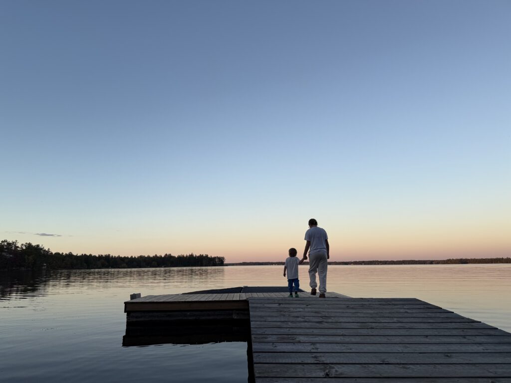 Two people standing at the end of a wooden dock overlooking a calm lake at sunset, with soft pastel sky reflections on the water.