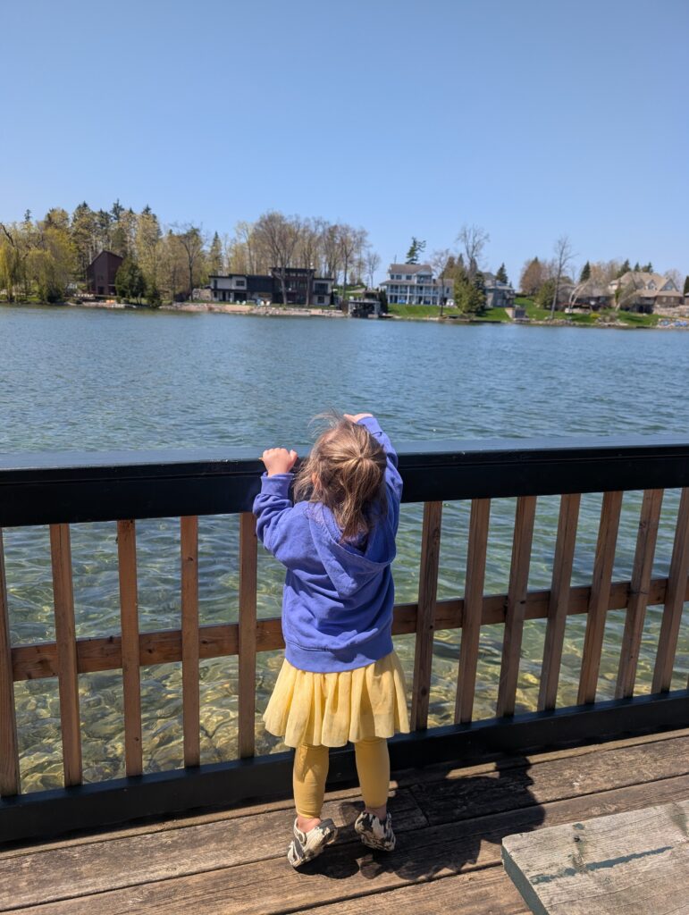 Young child standing at a wooden railing overlooking clear water and lakeside homes across Lake Couchiching