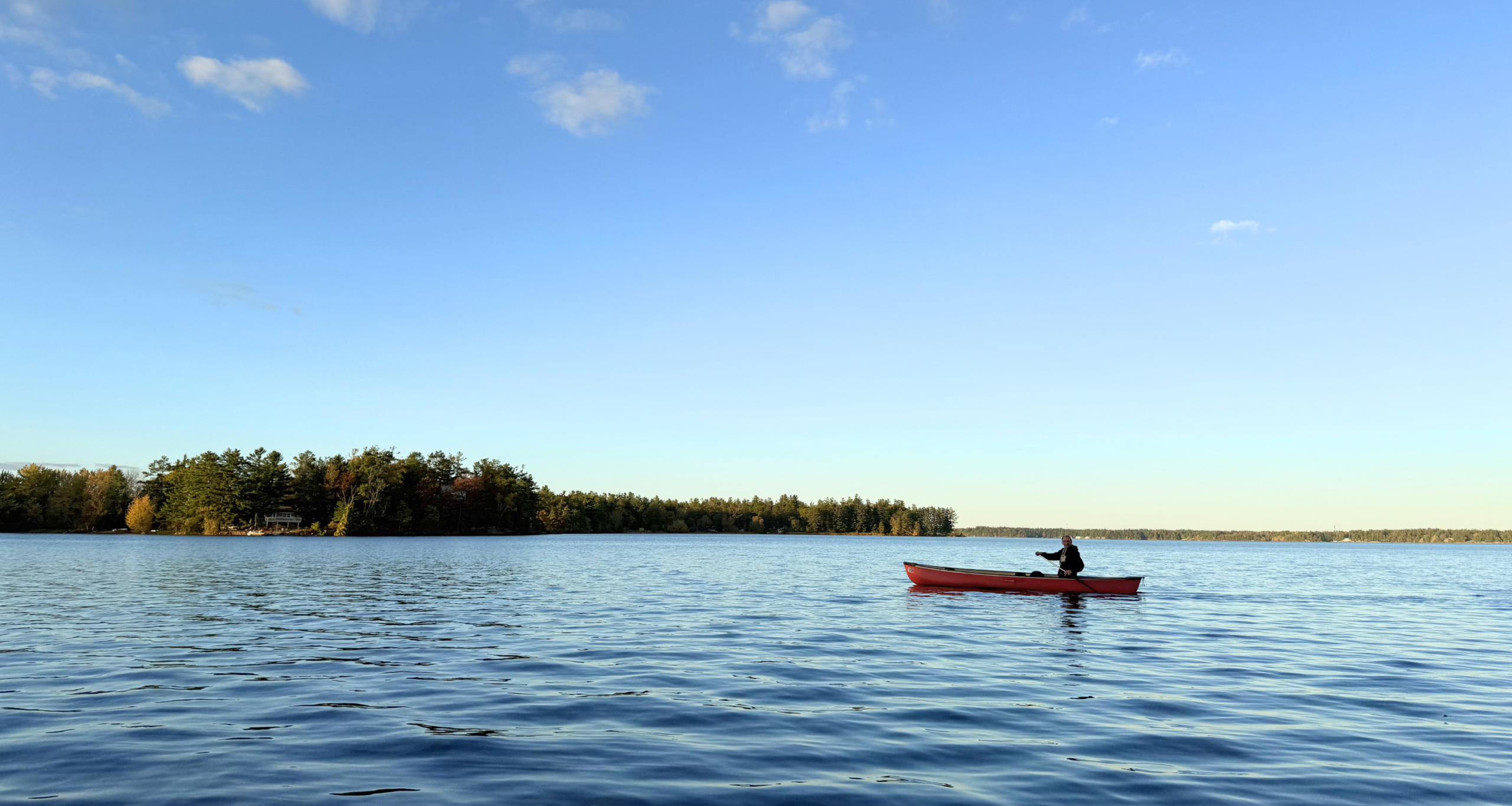 A person paddles a red canoe across the calm waters of Lake Couchiching under a wide blue sky, with a forested shoreline in the distance.