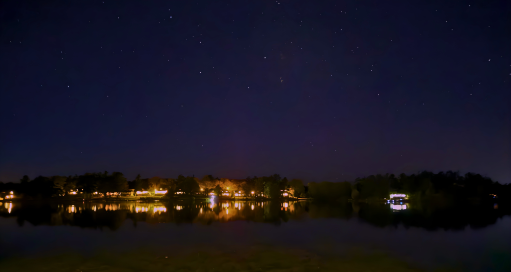 Stars fill the night sky above the calm waters of Lake Couchiching, where warm lights from homes along the shoreline reflect across the lake in the quiet darkness.